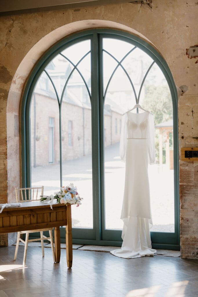 Bride’s wedding dress hanging by arched glass doors at Broxmouth Courtyard, soft natural light and rustic stone backdrop.