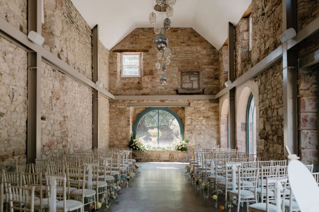 Wedding ceremony set up inside Broxmouth Courtyard with stone walls, arched window and aisle lined with white chairs.
