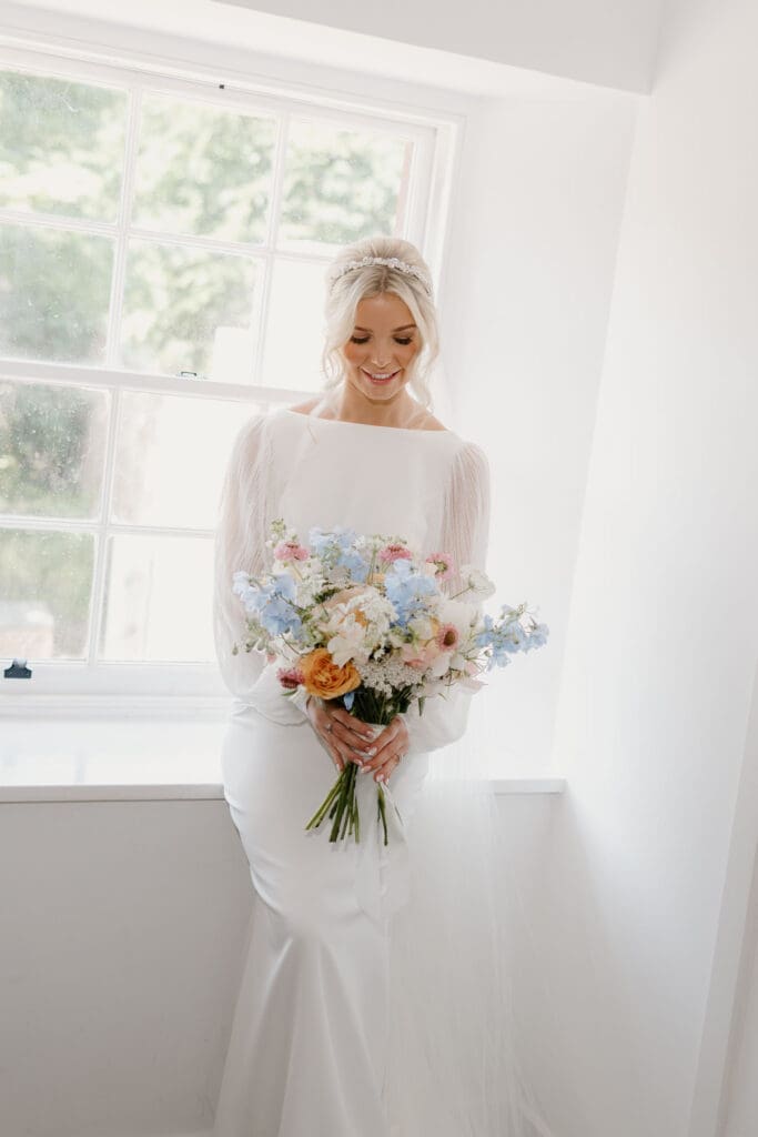 Bride standing by the window holding her bouquet in soft natural light.
