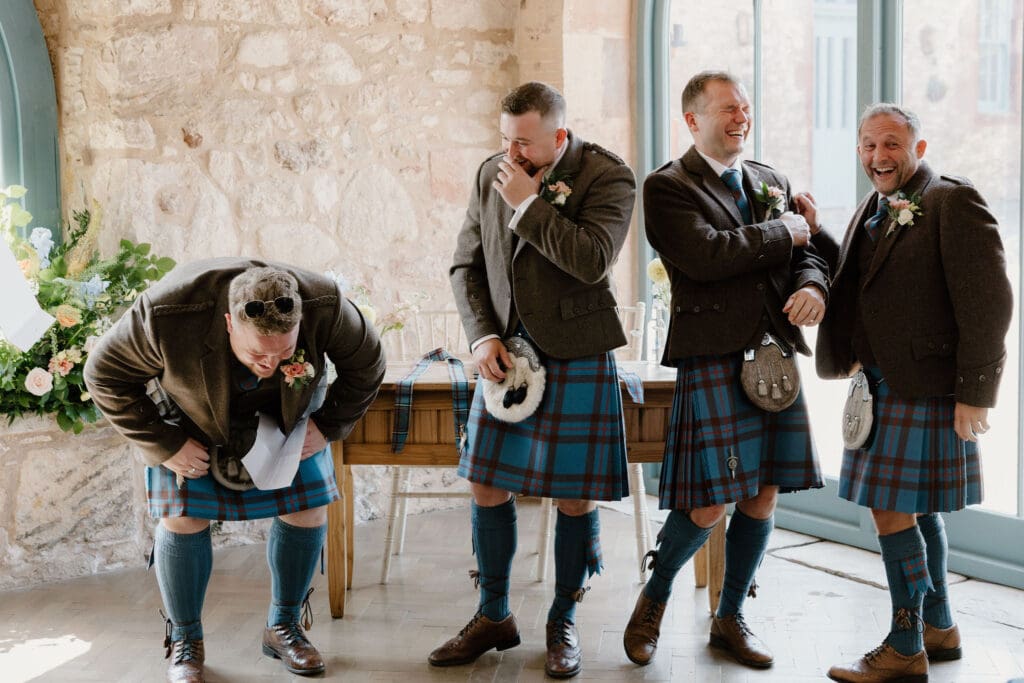 Groomsmen in traditional kilts laughing together during the wedding ceremony.