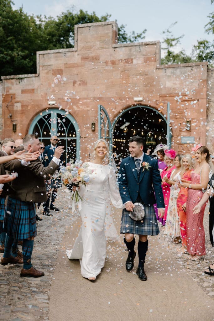 Bride and groom walking through a confetti throw outside Broxmouth Courtyard.