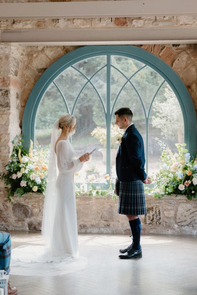 Bride and groom standing beneath the arched window during their wedding ceremony at Broxmouth Courtyard.