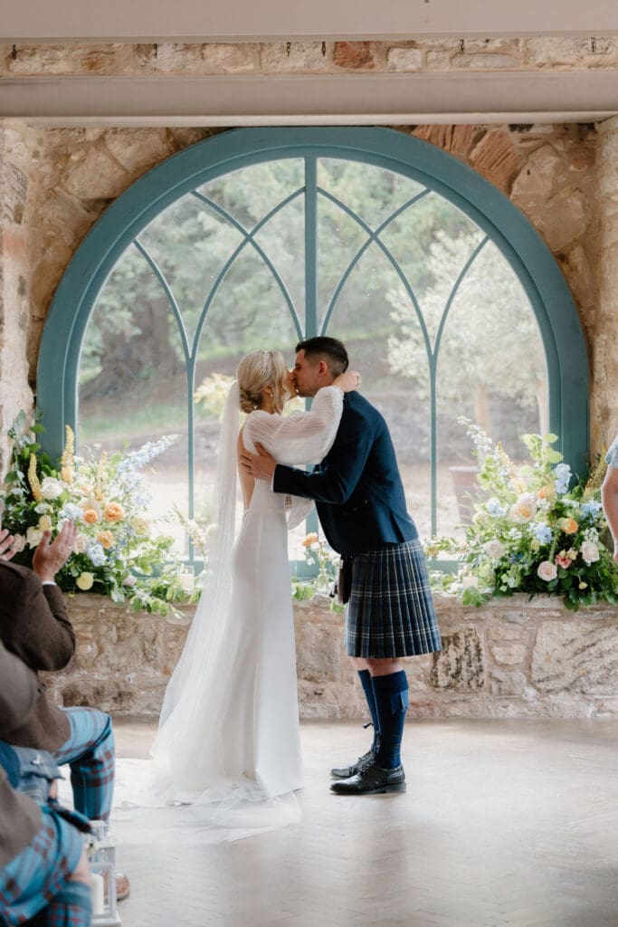 Bride and groom sharing their first kiss beneath the arched window at Broxmouth Courtyard.